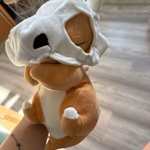 K&ouml;nnte beinhalten: Ein Cubone Pok&eacute;mon-Pl&uuml;schtier mit braunem K&ouml;rper und wei&szlig;em Sch&auml;delhelm. Das Spielzeug hat ein l&auml;chelndes Gesicht und wei&szlig;e Akzente an den Pfoten. Ein weiches Spielzeug f&uuml;r Pok&eacute;mon-Fans.