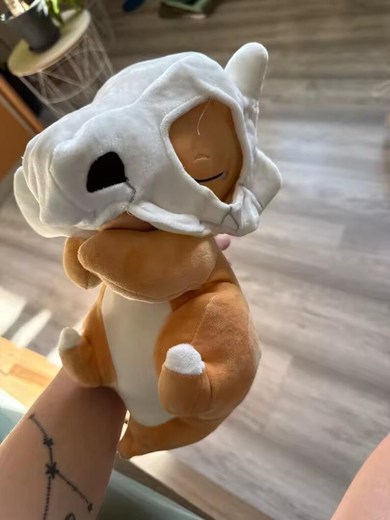 K&ouml;nnte beinhalten: Ein Cubone Pok&eacute;mon-Pl&uuml;schtier mit braunem K&ouml;rper und wei&szlig;em Sch&auml;delhelm. Das Spielzeug hat ein l&auml;chelndes Gesicht und wei&szlig;e Akzente an den Pfoten. Ein weiches Spielzeug f&uuml;r Pok&eacute;mon-Fans.