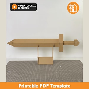 Puede incluir: Un modelo de espada de cartón color canela con punta, guarda y pomo, apoyado sobre un soporte rectangular. La imagen incluye un icono negro con el texto VIDEO TUTORIAL INCLUDED y un icono naranja con el texto PDF.