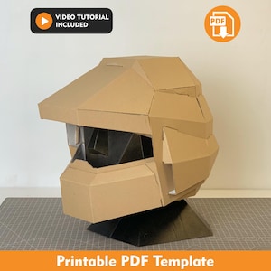 Puede incluir: Un casco de cartón color canela con un diseño geométrico y una amplia apertura de visera, montado sobre una base negra. La imagen incluye un icono negro con el texto VIDEO TUTORIAL INCLUDED y un icono naranja con el texto PDF, además de Printable PDF Template.