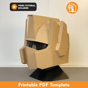 Op de afbeelding: Een bruine kartonnen helm met geometrische panelen en een vizier, inclusief een video-tutorial icoon en een PDF-download icoon. De helm staat op een zwarte standaard, met de tekst Printable PDF Template onderaan.