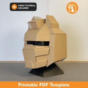 Puede incluir: Un modelo de casco de cartón color canela con paneles geométricos y una abertura para la visera, montado sobre una base negra. La imagen incluye iconos para un tutorial en video y una descarga en PDF, con el texto "VIDEO TUTORIAL INCLUDED" y "Printable PDF Template".