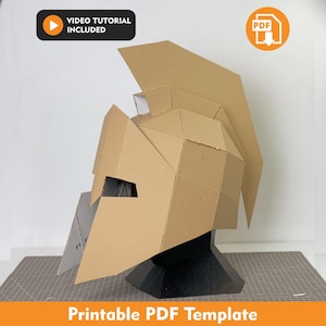 Peut inclure: Un casque de style spartiate en carton beige au design géométrique et bas polygone avec une haute crête. Le casque est présenté sur un support noir. L'image inclut des icônes pour un tutoriel vidéo et un téléchargement PDF, avec le texte Printable PDF Template.