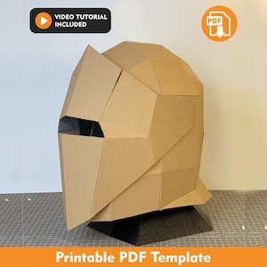 Puede incluir: Un casco de estilo geométrico low-poly construido con paneles de cartón color canela. El casco presenta una sección de visera puntiaguda y un interior oscuro visible a través de la abertura de los ojos. La imagen incluye iconos de tutorial en video y PDF.