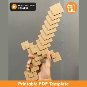 Puede incluir: Una espada pixelada construida con cuadrados de cartón marrón en capas, con un diseño en bloques. La imagen incluye un icono negro con el texto VIDEO TUTORIAL INCLUDED y un icono naranja con el texto PDF. El texto en la parte inferior dice Printable PDF Template.