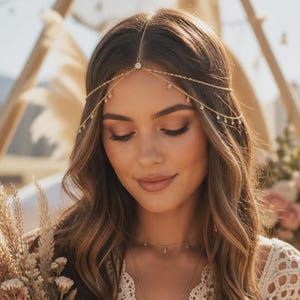 May include: A gold-toned headpiece featuring a delicate chain that rests across the forehead, accented with small, clear, round crystals. The design includes a central crystal drop and multiple smaller dangling crystals along the chain strands.
