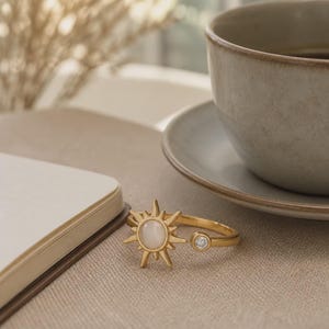 May include: A gold-toned open ring featuring a central sunburst motif with a smooth, round, milky white stone at its core. The opposite end of the band is finished with a single, small, clear, round-cut gemstone set in a simple gold bezel.