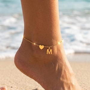 Könnte beinhalten: Goldfarbene Fußkettchen mit Herzanhänger und dem Buchstaben "M". Die Fußkette wird an einem Fuß an einem Sandstrand getragen, mit dem Meer im Hintergrund.
