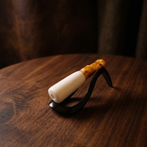 May include: A white and amber-colored smoking pipe with a black stand. The pipe has a cylindrical white body and a patterned amber mouthpiece. The black stand has a curved, minimalist design, resting on a dark wooden surface.