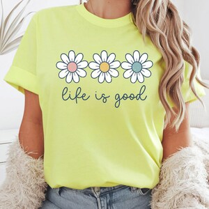 Op de afbeelding: Lichtgele t-shirt met drie madeliefjes met pastelroze, gele en blauwe centra. De woorden "life is good" staan in een sierlijk lettertype eronder. Het shirt heeft een casual stijl.