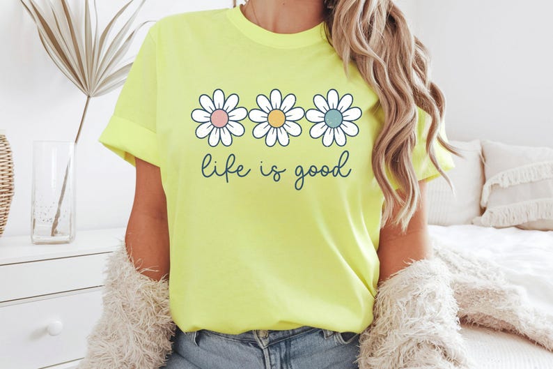 Op de afbeelding: Lichtgele t-shirt met drie madeliefjes met pastelroze, gele en blauwe centra. De woorden "life is good" staan in een sierlijk lettertype eronder. Het shirt heeft een casual stijl.