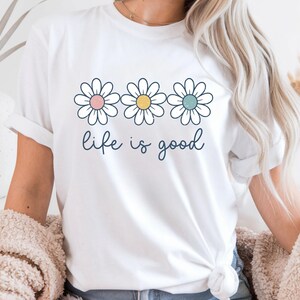 Op de afbeelding: Wit T-shirt met drie madeliefjes, elk met een verschillend gekleurd centrum: roze, geel en blauw. De woorden "life is good" zijn in een cursief lettertype onder de bloemen geschreven. Het shirt heeft opgerolde mouwen.