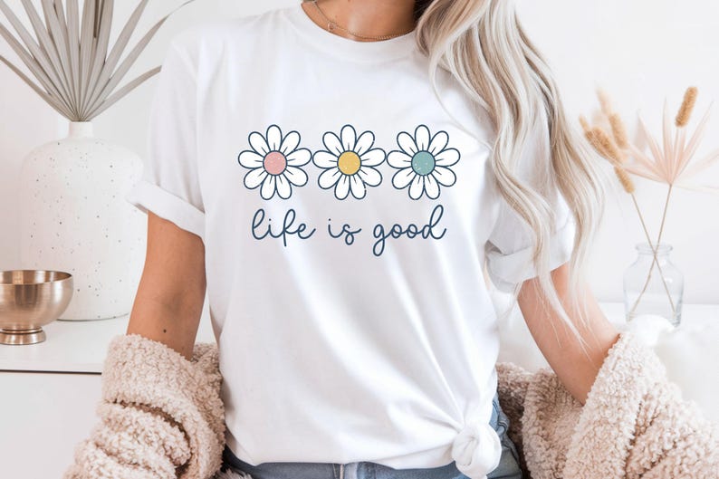 Op de afbeelding: Wit T-shirt met drie madeliefjes, elk met een verschillend gekleurd centrum: roze, geel en blauw. De woorden "life is good" zijn in een cursief lettertype onder de bloemen geschreven. Het shirt heeft opgerolde mouwen.