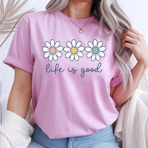 Op de afbeelding: Lichtroze T-shirt met opgerolde mouwen met drie madeliefjes met pastelkleurige centra en de tekst "life is good" in een scriptlettertype. Het T-shirt is lichtroze van kleur.