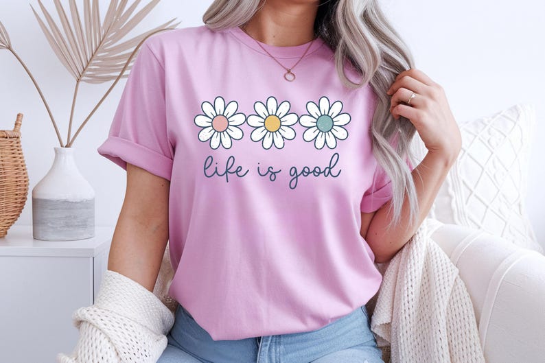 Op de afbeelding: Lichtroze T-shirt met opgerolde mouwen met drie madeliefjes met pastelkleurige centra en de tekst "life is good" in een scriptlettertype. Het T-shirt is lichtroze van kleur.