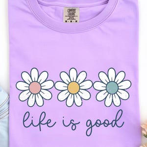 Op de afbeelding: Een lichtpaars T-shirt met drie madeliefjes, elk met pastelroze, gele en blauwe centra. De woorden "life is good" zijn in een cursief lettertype onder de bloemen geschreven. Het shirt is gemaakt van een zacht materiaal.