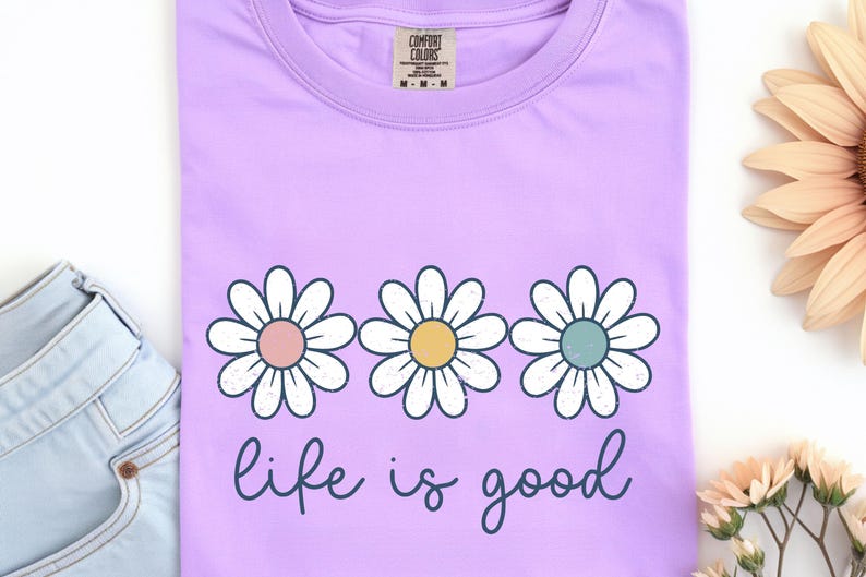 Op de afbeelding: Een lichtpaars T-shirt met drie madeliefjes, elk met pastelroze, gele en blauwe centra. De woorden "life is good" zijn in een cursief lettertype onder de bloemen geschreven. Het shirt is gemaakt van een zacht materiaal.