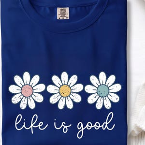 Op de afbeelding: Marineblauw T-shirt met drie madeliefbloemen met pastelroze, gele en blauwe centra. De tekst "life is good" staat in witte letters. Het T-shirt is gemaakt van zacht materiaal.