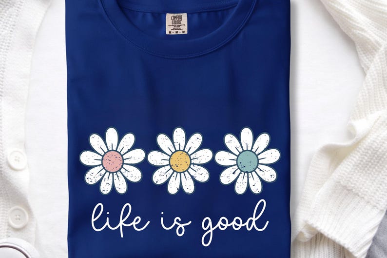 Op de afbeelding: Marineblauw T-shirt met drie madeliefbloemen met pastelroze, gele en blauwe centra. De tekst "life is good" staat in witte letters. Het T-shirt is gemaakt van zacht materiaal.
