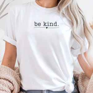 May include: White t-shirt with the phrase "be kind." printed in a simple, elegant font. The text is centered and underlined with a small heart symbol. The shirt has rolled-up sleeves.
