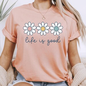Op de afbeelding: Perzikkleurig T-shirt met drie madeliefjes in wit, roze, geel en blauw. De zin "life is good" staat in een cursief lettertype onder de bloemen. Het shirt heeft opgerolde mouwen.