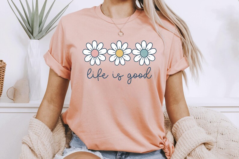 Op de afbeelding: Perzikkleurig T-shirt met drie madeliefjes in wit, roze, geel en blauw. De zin "life is good" staat in een cursief lettertype onder de bloemen. Het shirt heeft opgerolde mouwen.