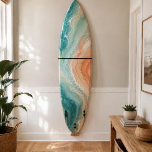 Può includere: Una tavola da surf montata verticalmente su una parete, caratterizzata da un design artistico astratto in resina che imita le onde dell'oceano che si infrangono su una spiaggia sabbiosa con sfumature vorticose di turchese, blu, schiuma bianca e pesca.