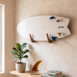 Op de afbeelding: Een wit surfboard met vier vinnen, horizontaal aan de muur bevestigd met twee houten beugels. Het board heeft een visstaartontwerp en een klein rond logo. Op een houten plank eronder staan een plant, een enkele vin en een stapel boeken.