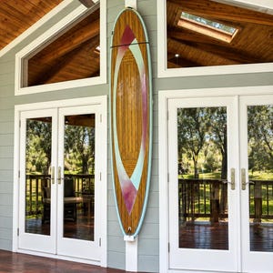 Può includere: Una tavola da surf verticale con finitura in venature di legno naturale, caratterizzata da un design geometrico centrale nei toni dell'azzurro e del magenta intenso. La tavola è montata su una parete tra due porte bianche vetrate.
