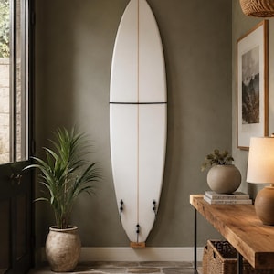 Puede incluir: Una tabla de surf blanca con un alma central de madera y tres quillas negras montada verticalmente en una pared. La tabla está asegurada por una correa negra y descansa sobre una pequeña base de madera, con punta afilada y cola redondeada.