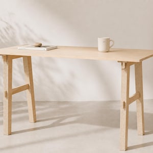 May include: A light-colored wooden desk with a rectangular top and angled legs. A white mug and a book are on the desk. A surfboard leans against the wall. The desk is approximately 60 inches long.