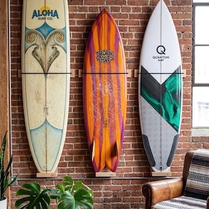 Könnte beinhalten: Drei Surfbretter an einer Ziegelwand. Das linke Brett ist cremefarben mit blauen und goldenen Akzenten und dem Text Aloha Surf Co. Das mittlere Brett ist orange und lila mit dem Text Tropic Waves. Das rechte Brett ist weiß, schwarz und grün mit dem Text Quantum Surf.