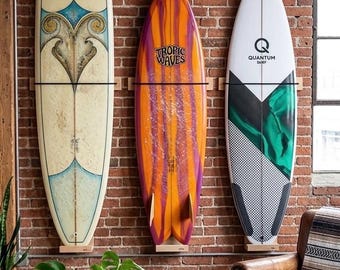 May include: Three surfboards mounted on a brick wall. The left board is cream with blue and gold accents and the text Aloha Surf Co. The center board is orange and purple with the text Tropic Waves. The right board is white, black, and green with the text Quantum Surf.