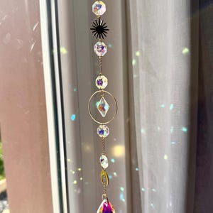 Op de afbeelding: Een decoratieve zonnevanger met een gouden ketting en iriserende kristallen. De zonnevanger heeft een ster, een cirkel en een druppelvormig kristal. De kristallen reflecteren licht en creëren een regenboogeffect.