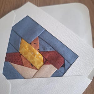 Könnte beinhalten: Handgefertigte Grußkarte mit einem farbenfrohen geometrischen Design. Das Design der Karte umfasst blaue, gelbe, rote und beige Stoffstücke. Die Karte befindet sich in einem weißen Umschlag, ideal für eine besondere Nachricht.