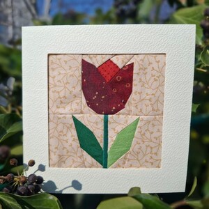 Könnte beinhalten: Ein gerahmtes Stoffkunstwerk mit einer gesteppten Tulpe. Die Tulpe hat eine weinrote Blüte mit Goldpunkten, grüne Blätter und einen grünen Stiel. Der Hintergrund ist ein beiger Stoff mit einem goldenen Blumenmuster. Der Rahmen ist weiß.