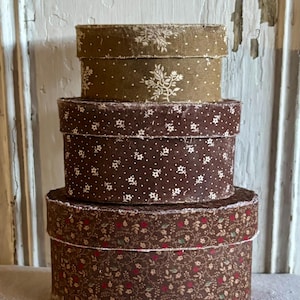 May include: A stack of three round fabric-covered boxes. The top box is olive green with a white floral pattern. The middle box is dark brown with a white floral print. The bottom box is brown with a small red and green floral pattern.