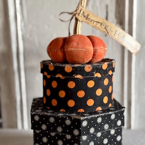 May include: A decorative orange fabric pumpkin sits atop a round black box with orange polka dots, which rests on a square black box with white snowflake patterns. A tan paper tag tied to the pumpkin stem reads Merry Olde Halloween.