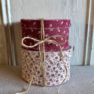 May include: Two round fabric boxes stacked on top of each other, tied with twine. The top box is burgundy with white floral accents. The bottom box is white with burgundy floral accents. The twine is tied in a bow.