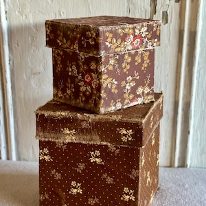 May include: Two stacked square boxes with lids, covered in brown fabric. The top box features a dense floral pattern in red, white, and gold. The bottom box displays a smaller, repeating gold floral and dot pattern with frayed fabric edges.