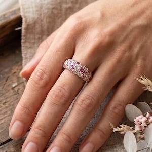 May include: A silver-toned band ring featuring a pavé setting with alternating round pink and clear stones. The ring is worn on a finger, positioned near a small bouquet of dried pink roses and eucalyptus leaves on a rustic wooden surface.