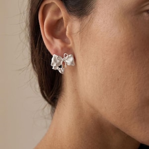 May include: A silver-toned stud earring shaped like a small bow with a matte finish. The earring features a central knot detail with two loops and two trailing ribbon ends, securely fastened to the earlobe with a standard post and backing mechanism.