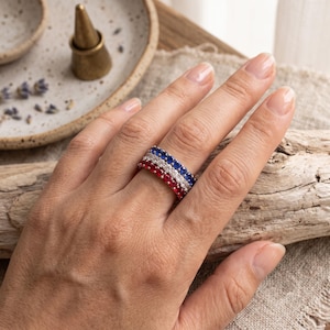 May include: A stack of three eternity bands worn on a finger, featuring a row of round-cut blue stones, a row of round-cut clear stones, and a row of round-cut red stones, each set in a metallic band with a polished finish.