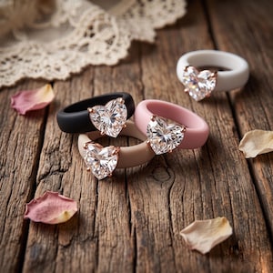 May include: Three silicone rings featuring heart-shaped clear faceted stones set in rose gold-toned metal prongs. The bands are solid matte colors in black, soft pink, and white, each showcasing a prominent central heart-shaped gemstone.