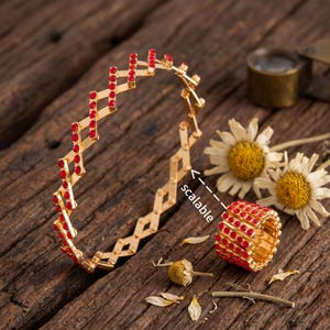 May include: A gold-toned expandable bracelet and a matching ring, both featuring a lattice design set with small red crystals. The word scalable is printed with an arrow pointing to the bracelet. The jewelry is displayed with white flowers.