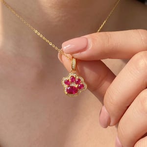 May include: A gold necklace featuring a pendant shaped like an animal paw print. The pendant is set with five oval-cut red rubies and accented with small white pavé stones along the border and the bail, suspended from a delicate gold chain.