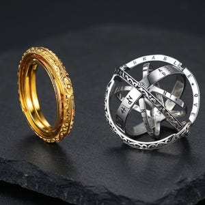 Op de afbeelding: Een goudkleurige ring met ingewikkelde krulpatronen naast een zilverkleurige astronomische bolring. De zilveren bol is voorzien van gegraveerde letters en symbolen op de roterende banden, die kunnen worden uitgevouwen tot een complexe bolvorm.