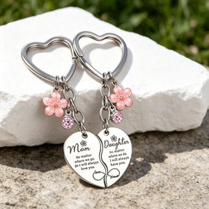 May include: Two silver heart-shaped keychains, one reading "Mom" and the other "Daughter." Each has a pink flower charm and a pink crystal. The keychains are connected and feature the text "No matter where we go, I will always love you."