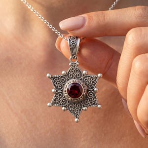 May include: A silver pendant necklace featuring a star-shaped design with intricate filigree detailing. The pendant has a central, round, deep red gemstone. The necklace is on a delicate silver chain.