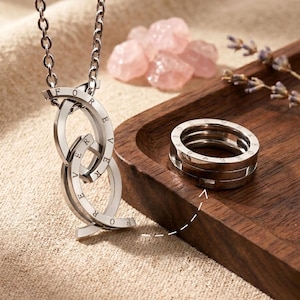 Puede incluir: Un collar de metal plateado con dos colgantes ovalados entrelazados grabados con la palabra FOREVER, suspendidos de una cadena a juego, acompañado de un anillo plateado con el mismo grabado, ambos con un acabado metálico pulido.